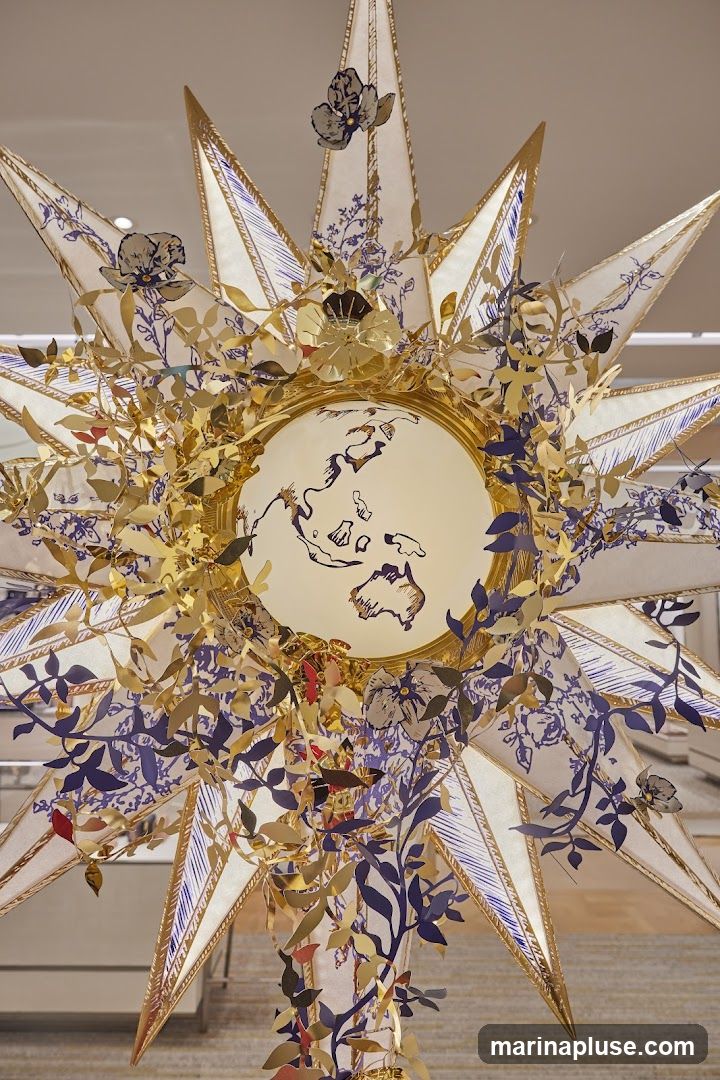 A close-up view of an elaborate sunburst-shaped decoration, likely a window display or interior decor. The central part features a circular frame with a depiction of a world map, possibly of Asia and Australia. The sunburst itself is adorned with numerous gold leaf cutouts resembling foliage and flowers, interspersed with dark blue floral and vine patterns printed on white fabric panels. The overall aesthetic is intricate and artistic, suggesting a high-end retail or event setting.