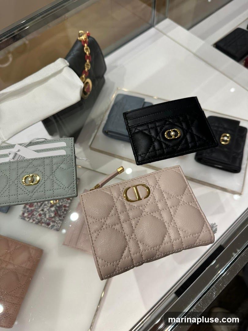 A display of various quilted leather accessories, including cardholders and small wallets, in pastel pink, light blue, and black. Each item features the iconic CD logo and a quilted pattern. A black handbag with a red and gold chain is also visible in the background.