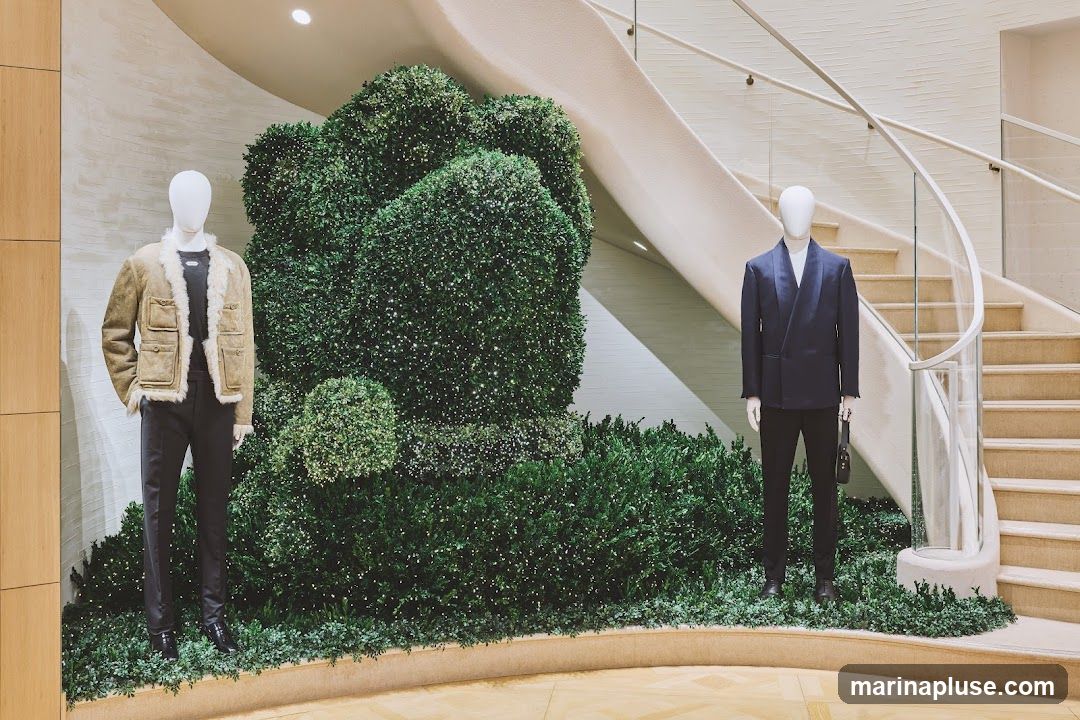 Two mannequins are displayed in a store. The mannequin on the left wears a tan suede jacket with shearling trim over a dark t-shirt and dark pants. The mannequin on the right wears a dark blue satin-lapel jacket over a white shirt and dark pants, and holds a small black handbag. They are positioned in front of a large, sculpted green hedge adorned with small white lights. A curved staircase with glass railings is visible in the background.