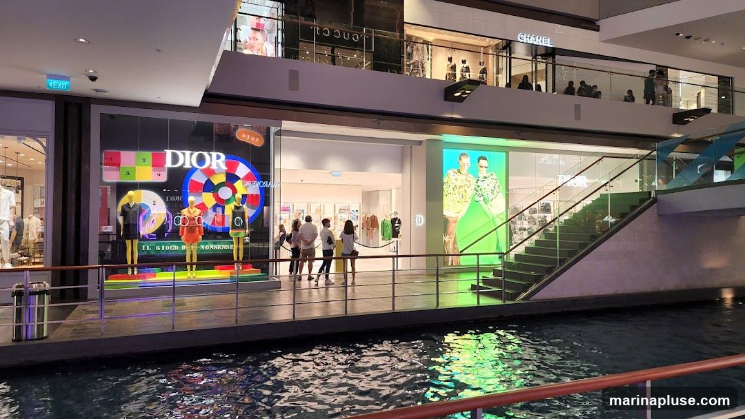 The storefront of a Dior store is visible, featuring mannequins dressed in colorful outfits and a large, circular graphic with the Dior logo. The store's interior is visible through the glass, displaying handbags and clothing. To the right of the Dior store, a large digital screen shows two models. Above, the Chanel store is also visible.