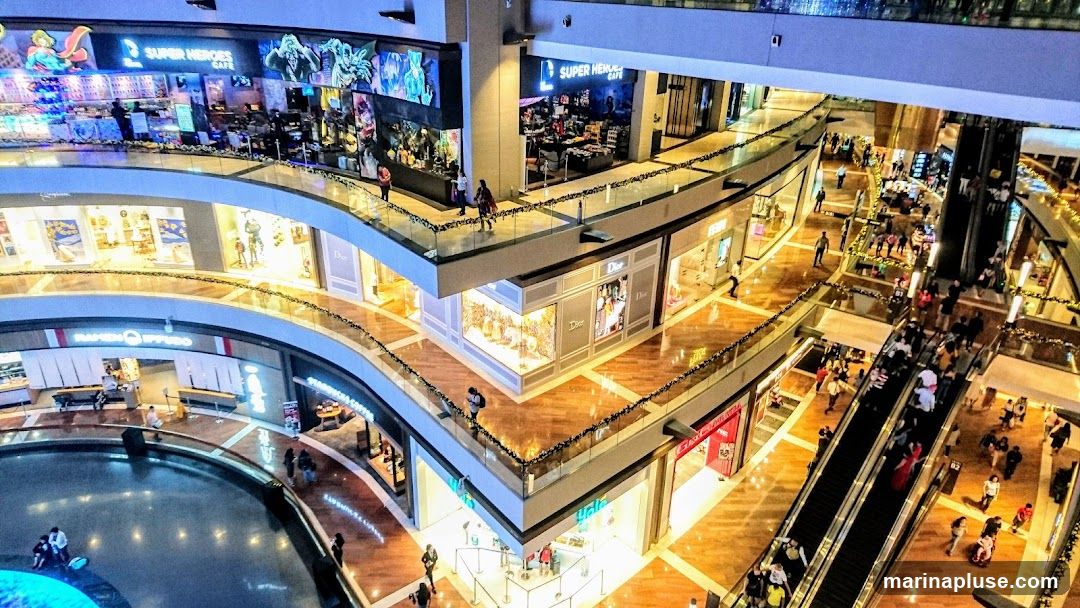 An aerial view of the interior of a bustling shopping mall. Multiple levels are visible, with shops lining the curved walkways. People are seen walking, shopping, and riding escalators. Prominent signage for 'Super Heroes Cafe' and 'Dior' are visible. A water feature is present on a lower level.
