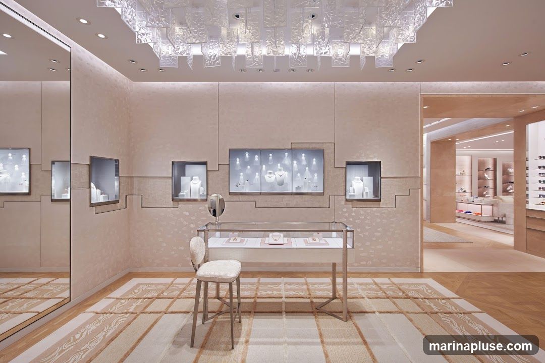 The interior of a high-end jewelry store is depicted, featuring elegant display cases filled with necklaces and other jewelry. The walls are textured and a large mirror reflects the space. A plush stool sits in front of a glass display table. The ceiling has a decorative, modern light fixture. In the background, another section of the store is visible with more displays and shelving.