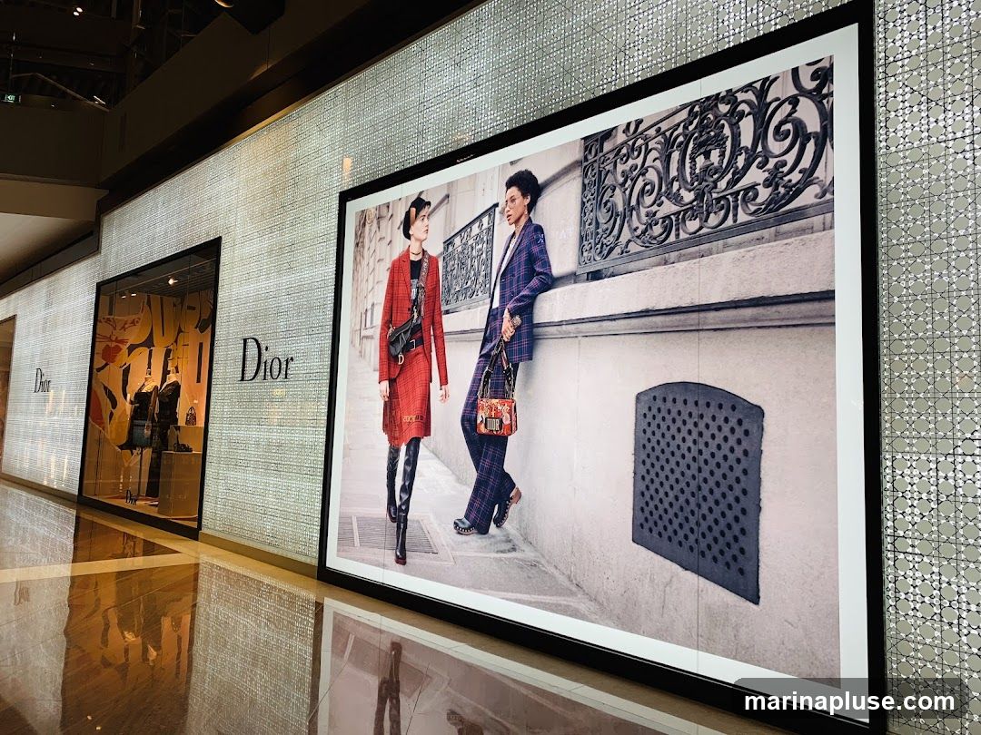 A wide shot of a Dior store interior and exterior display. The left side shows a store window with mannequins dressed in clothing and handbags. The Dior logo is visible on the wall next to the window. To the right of the window is a large framed advertisement featuring two models posing outdoors. The advertisement also displays the Dior logo. The floor is highly reflective, mirroring the store and advertisement.