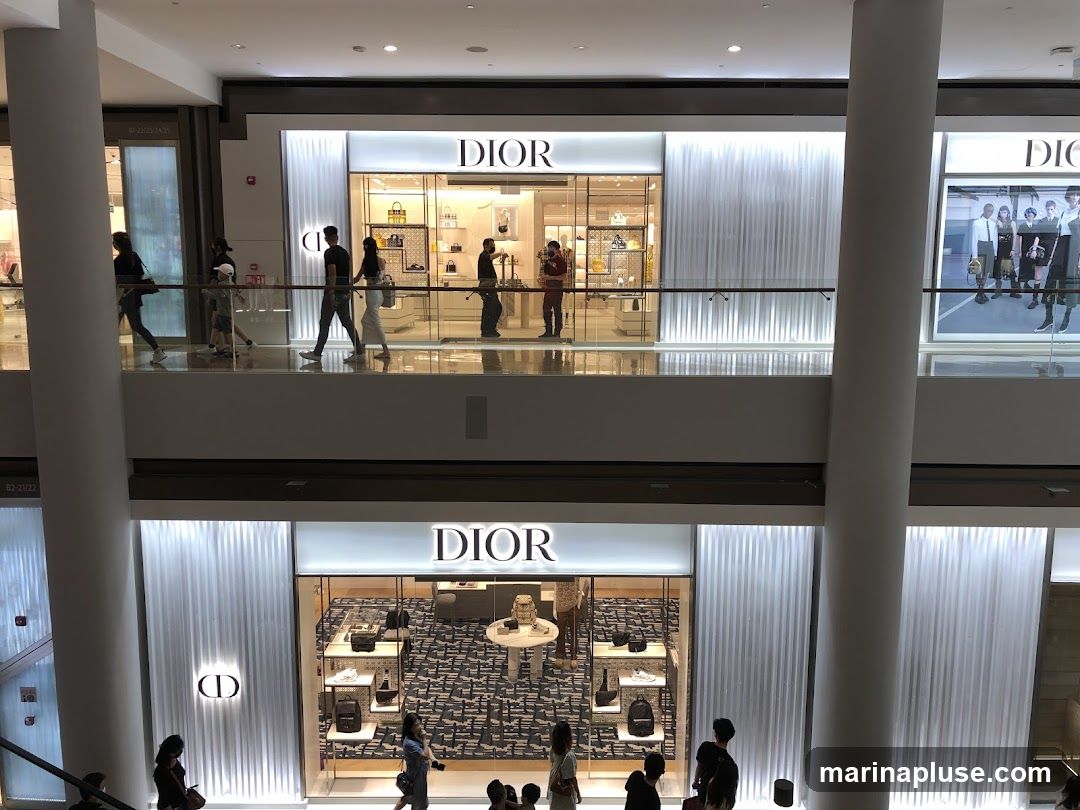 A view of two Dior storefronts on different levels of a mall. The upper storefront displays handbags and accessories. The lower storefront showcases handbags, shoes, and other accessories on shelves and a central display table. People are seen walking on the upper level walkway and on the lower level in front of the store.