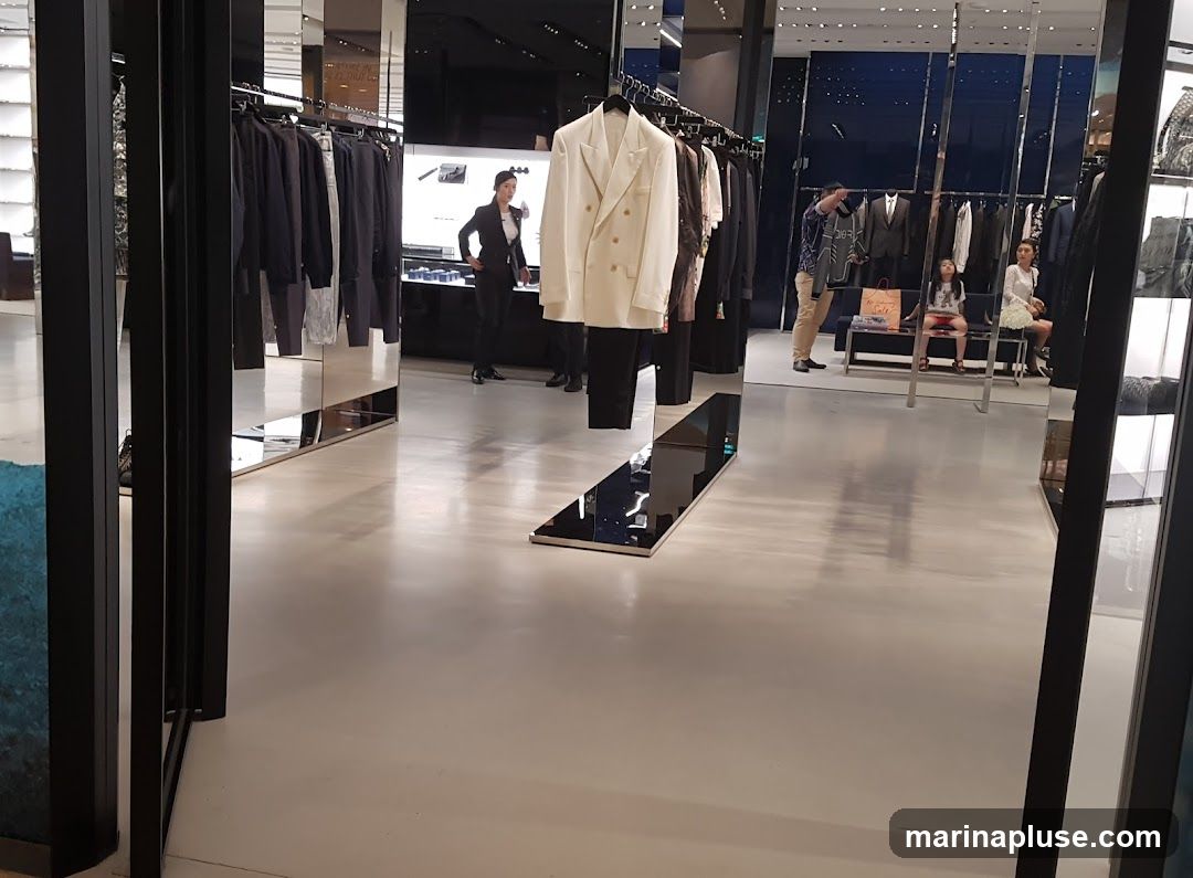 A view inside a high-end clothing store, showcasing racks of apparel and mannequins. The store features a modern design with polished floors and mirrored surfaces. Several customers and staff members are visible browsing the merchandise and interacting with each other. A white double-breasted blazer is prominently displayed on a hanger in the foreground.
