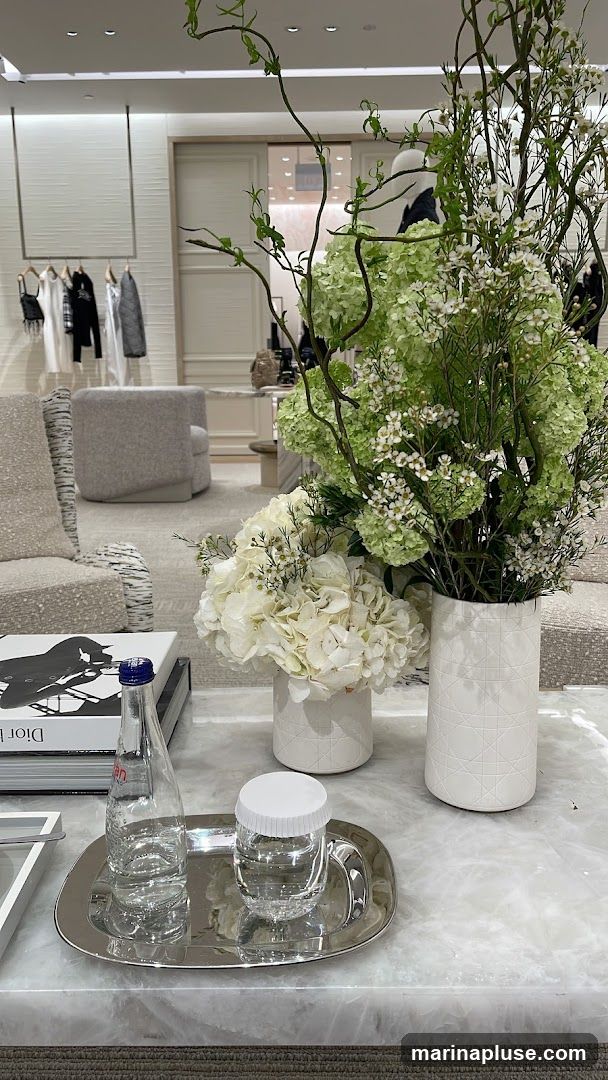 A coffee table in a store displays a book with the Dior logo, a bottle of Evian water, and a glass of water. Two white vases hold arrangements of white and green flowers. In the background, clothing is displayed on racks, and seating areas are visible.