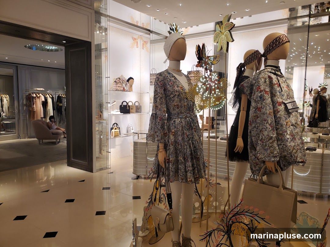 A well-lit interior of a luxury fashion store, likely Dior, with mannequins dressed in floral print clothing and accessories. The display features handbags, dresses, and outerwear. In the background, racks of clothing and a seating area are visible. The store's polished floor reflects the bright lighting and displays.