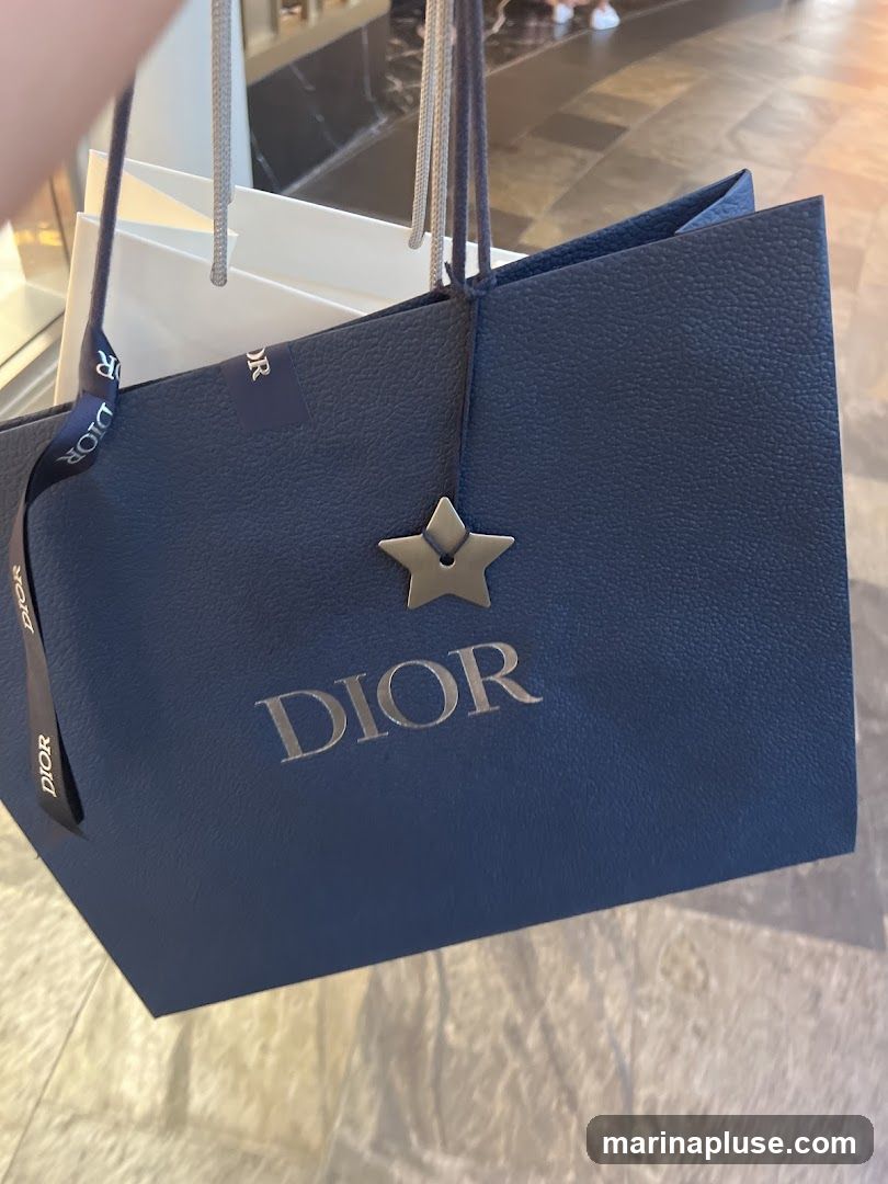 A close-up shot of a dark blue Dior shopping bag with the brand name embossed in silver. A silver star charm hangs from one of the bag's handles. Another white shopping bag is visible behind it. The background appears to be a store interior with tiled flooring and blurred figures of people.