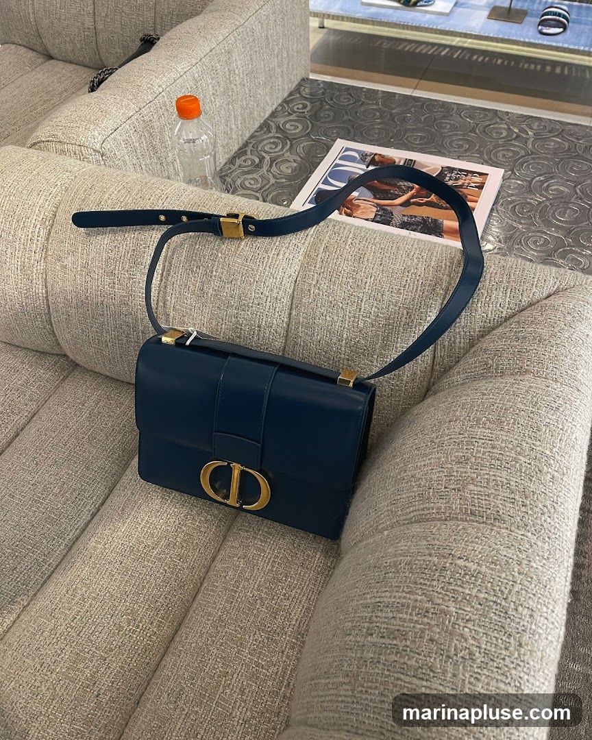 A dark blue Dior handbag with gold hardware rests on a textured grey sofa. A magazine with the Dior logo is visible on a patterned table next to the sofa, along with a bottle of water.