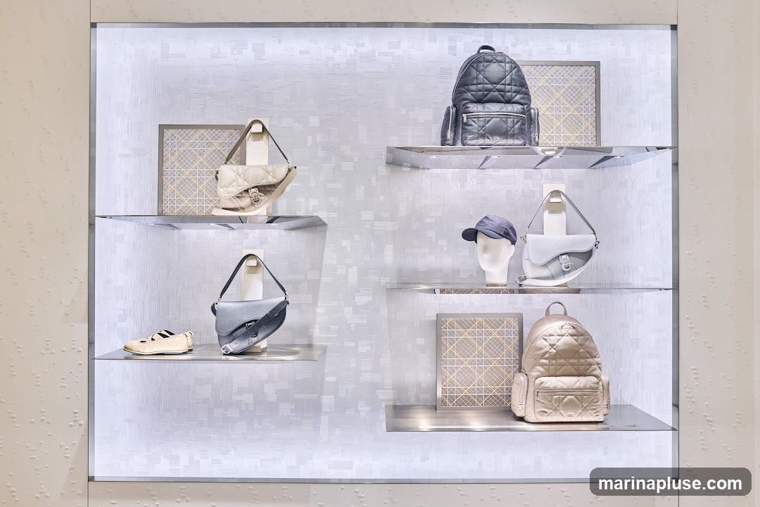 A well-lit store window display featuring various Dior handbags, backpacks, and shoes. The display is organized on multiple shelves with a textured, light-colored background. Items include a beige saddle bag, a grey saddle bag, a pair of beige flats, a grey backpack, a white mannequin head wearing a grey cap, a light blue saddle bag, and a light pink backpack. Decorative framed squares are also placed on some shelves.