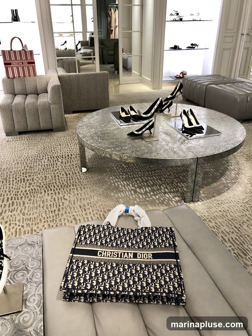 A Christian Dior tote bag with the brand's signature monogram pattern is displayed on a gray tufted bench. Next to it, on a circular metallic table, are several pairs of elegant high-heeled shoes. The background shows a well-lit boutique interior with shelves displaying more footwear and clothing.