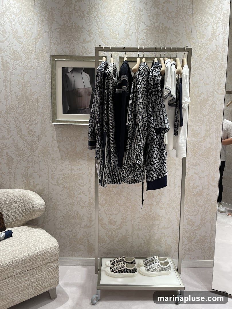 A clothing rack with Dior branded apparel, including jackets and shirts, is displayed in a store. Below the rack, a pair of Dior sneakers are showcased. A textured armchair is visible to the left, and a framed image of a mannequin is on the wall. The store features ornate wallpaper and a large mirror on the right side.