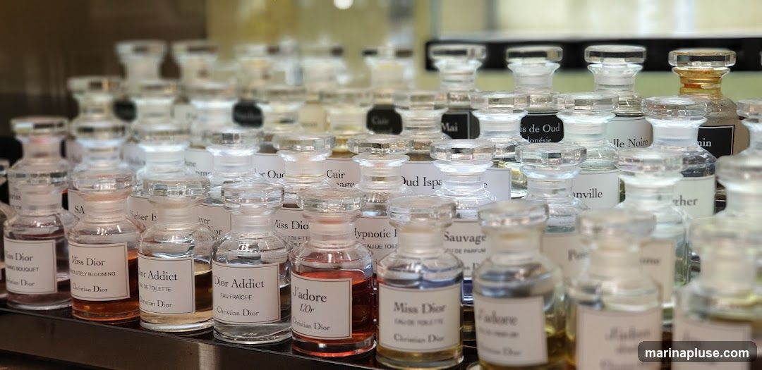 A display of numerous small glass perfume bottles with clear stoppers, arranged in rows. The labels on the bottles indicate various fragrances, including "Miss Dior," "Dior Addict," and "J'adore," along with brand names like "Christian Dior." The bottles contain liquids of different colors, from clear to amber.
