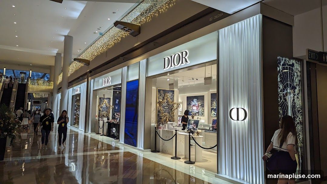 The exterior of a Dior store in a shopping mall. The store has large glass windows displaying merchandise and illuminated signage with the Dior logo. The facade is modern and sleek, with vertical white ribbed panels and decorative lighting. People are seen walking in the mall outside the store.