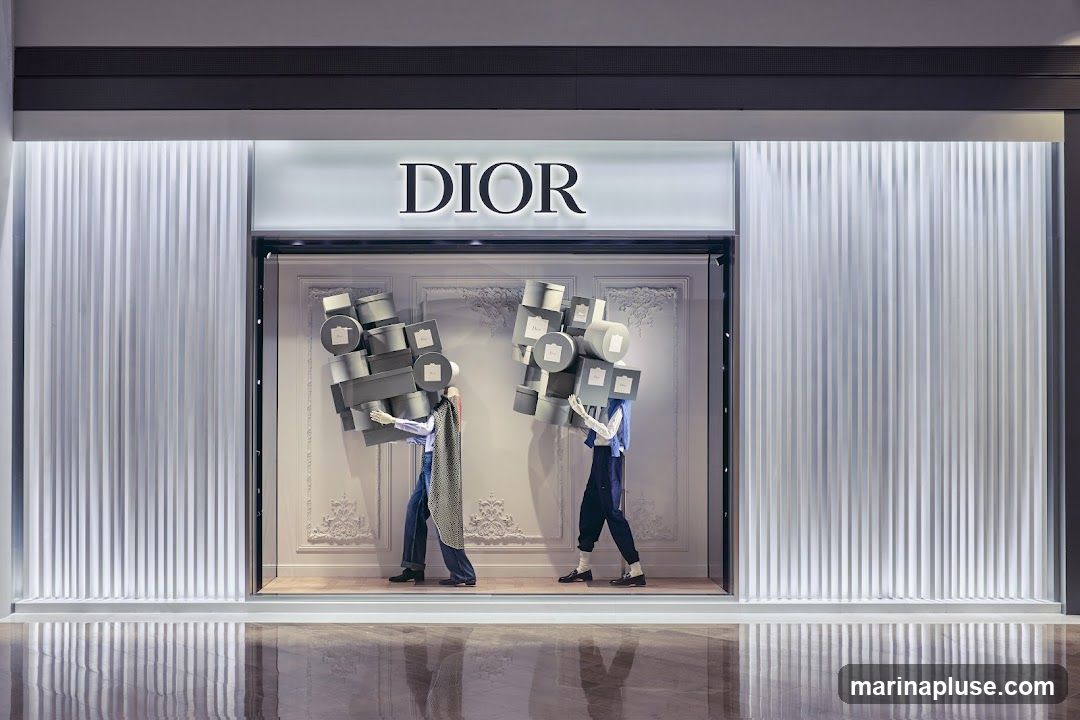 The exterior of a Dior store features a large, illuminated sign with the brand name. The storefront window display showcases two mannequins, each carrying a large, precarious stack of Dior-branded boxes. The mannequins are dressed in stylish outfits, and the background of the display is ornate and decorative. The overall presentation is elegant and eye-catching, typical of luxury retail window displays.