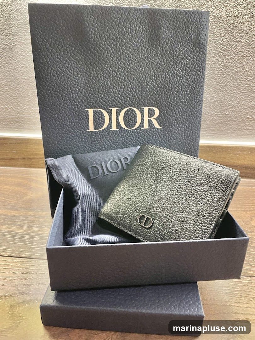 A black pebbled leather Dior wallet is displayed in its original packaging, which includes a dark blue textured box and a matching shopping bag with the Dior logo. A dark grey fabric pouch with the Dior logo is also visible, partially tucked under the wallet.