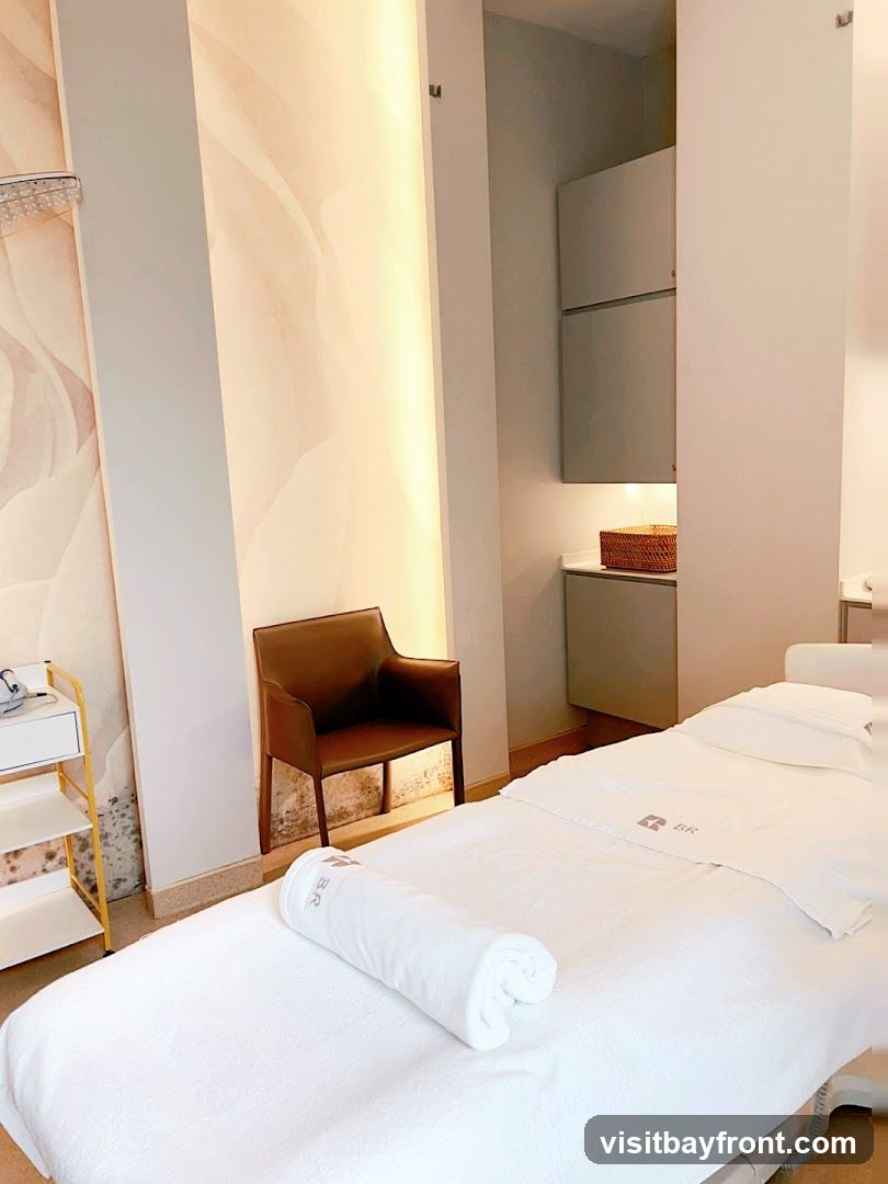 A clean and serene spa treatment room with a massage bed covered in white linens, a rolled towel, and a brown leather chair. The room features soft lighting, a decorative wall mural, and storage cabinets.