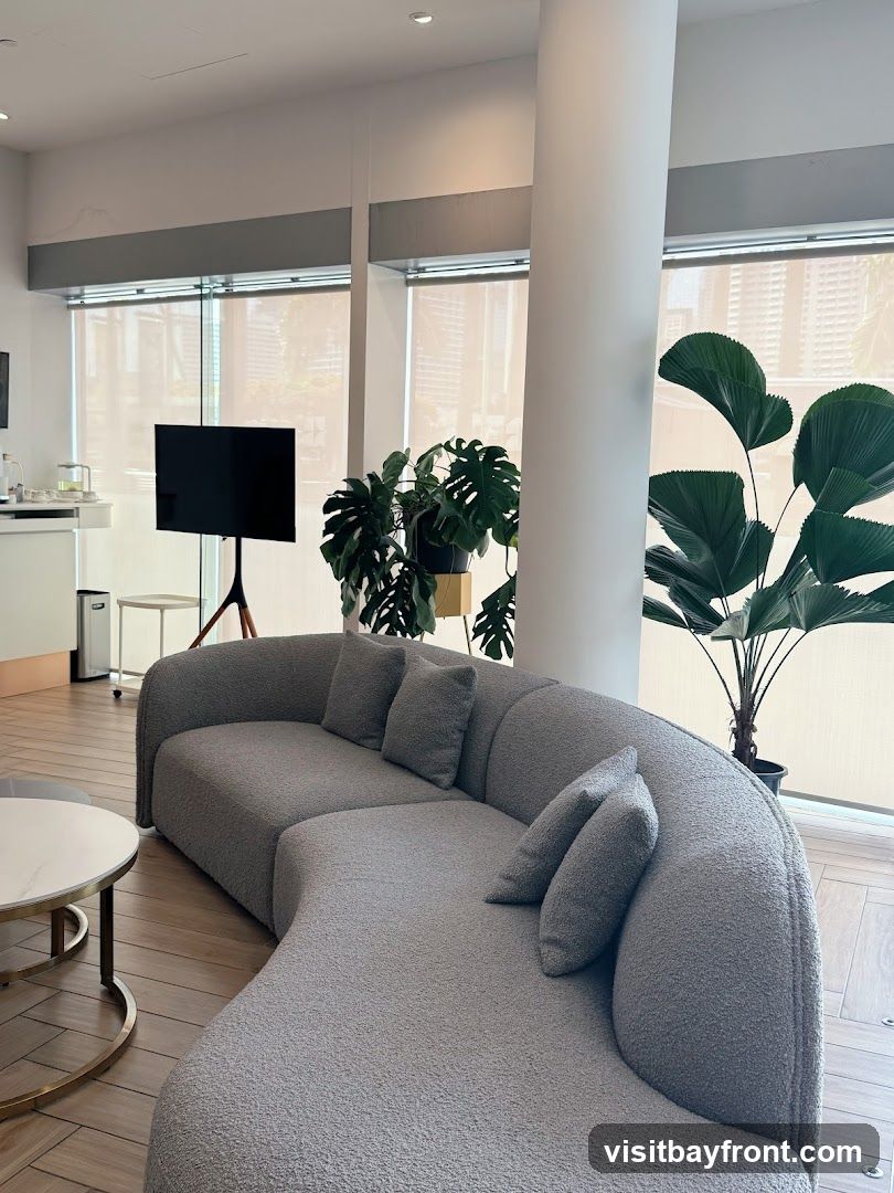 A contemporary lounge area with a large, curved grey sofa, plush cushions, and a round marble-topped coffee table with a gold base. Large windows with blinds allow natural light to fill the space, illuminating several potted plants, including a Monstera and a fan palm. A black television on a tripod stand is visible in the background, along with a white counter and a silver trash can. The flooring is light-colored wood.