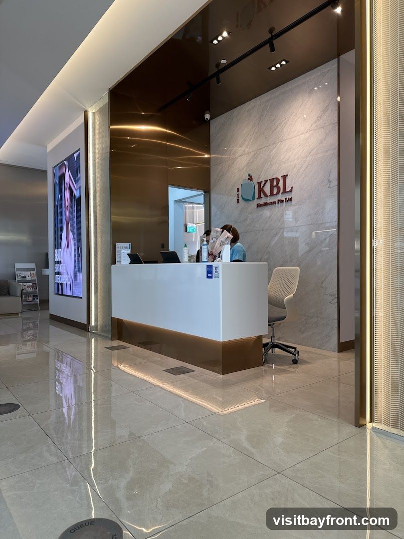 A modern reception area with a white desk, marble walls, and a large digital display. Two receptionists are visible behind the desk. The KBL Healthcare Pte Ltd logo is displayed on the wall.
