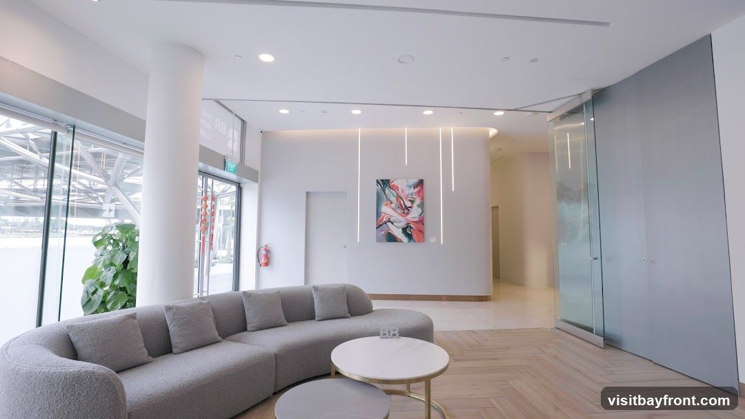 A contemporary lobby features a curved grey sofa with plush cushions, two round coffee tables, and a large potted plant near a floor-to-ceiling window. The walls are white, with a modern abstract painting displayed prominently. Vertical LED lights add a sleek touch to the decor. A fire extinguisher is visible on the left, and a glass-enclosed doorway is on the right. The flooring is a light-colored herringbone pattern.