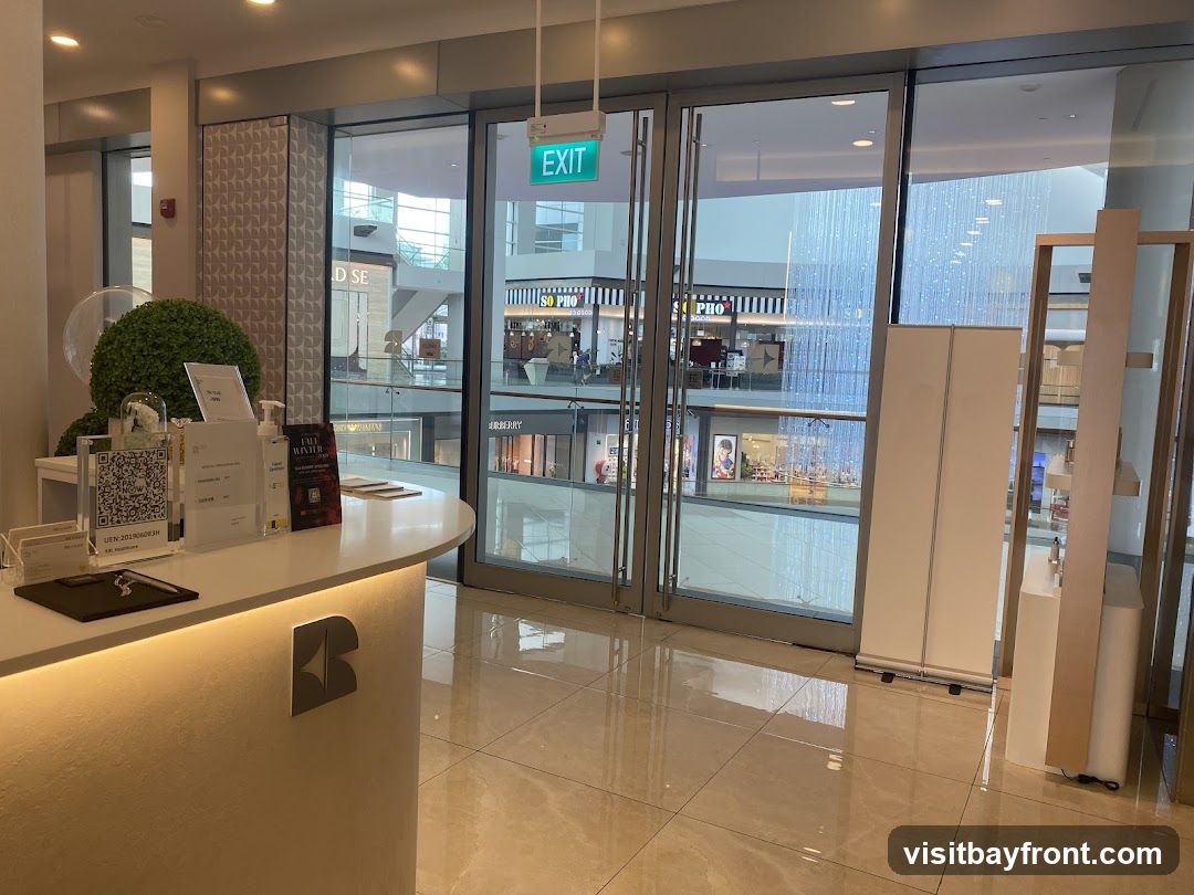 A reception desk with a logo is visible in the foreground. Behind the desk, large glass doors offer a view into a shopping mall with various storefronts and decorative lights. An 'EXIT' sign is illuminated above the doors.