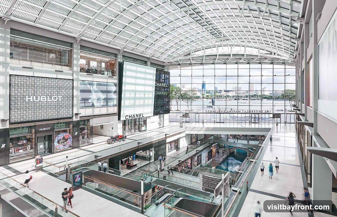 The image shows the interior of a multi-level shopping mall. Large digital screens display advertisements for brands like Hublot and Chanel. Below these screens are the storefronts of various luxury brands. The mall features an indoor canal with boats, and escalators connect the different levels. People are seen walking around the mall. The overall impression is one of a high-end, spacious, and contemporary retail environment.