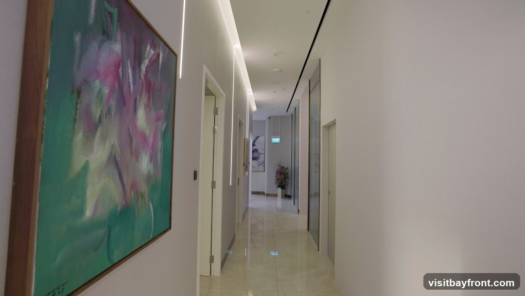 A long, well-lit hallway with a polished tiled floor. On the left wall, a large abstract painting with green and pink hues is displayed. The hallway features recessed lighting along the ceiling and vertical accent lights on the walls. Several doors line both sides of the hallway. In the distance, an illuminated green 'EXIT' sign is visible, along with a potted plant.