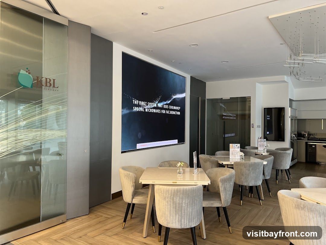 A contemporary waiting area with plush grey chairs and white tables. A large digital screen displays text about "COOLWAVES" for fat reduction. The space features a glass-enclosed area with a logo visible on the glass, and a small kitchen or service area in the background.