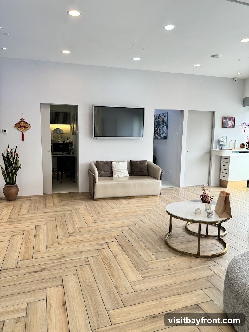 A waiting area with a beige sofa, a coffee table with a marble top, and a large television mounted on the wall. The floor is made of light-colored wood laid in a herringbone pattern. There is a plant in a pot on the left side of the room and artwork on the walls.