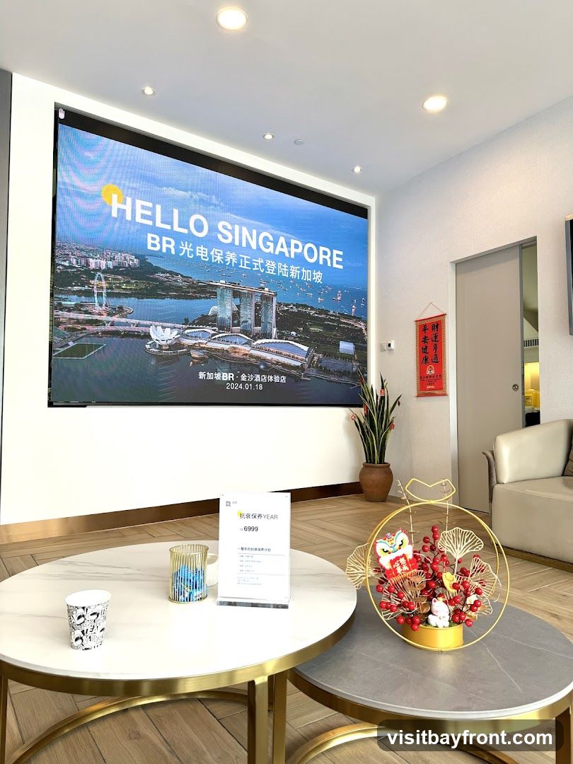 The image shows the interior of a modern space with a large LED screen displaying "HELLO SINGAPORE" and an image of the Singapore skyline. Below the main display, text in Chinese indicates the opening of a BR store in Singapore. In the foreground, there are two round coffee tables. The left table holds a paper cup and a decorative item. The right table has a small stand with text and pricing information, and a festive decorative arrangement with a lion dance figure and a lucky cat. A sofa is visible in the background.