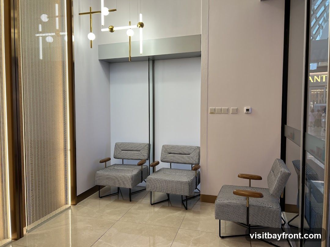 A waiting area with three chairs upholstered in a black and white houndstooth pattern, each with wooden armrests. The chairs are arranged in a row against a plain white wall. To the left, a decorative screen with a herringbone pattern is illuminated by vertical LED lights. Above the chairs, a modern chandelier with gold and white elements hangs from the ceiling. Light-colored tiled flooring reflects the ambient light. In the background, through a glass partition, a glimpse of a shopping area with signage is visible.