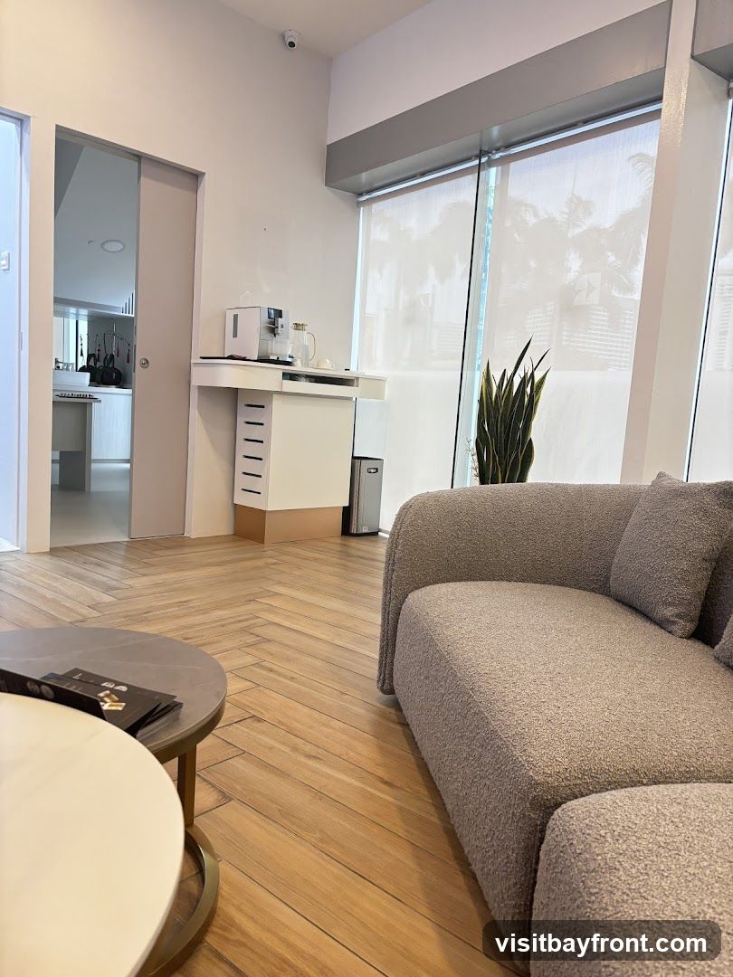 A contemporary waiting area with a plush grey sofa, two round coffee tables, and a sleek white console with a coffee maker and carafe. A tall potted plant adds a touch of greenery near the large window with sheer blinds. The floor is made of light-colored wooden planks laid in a herringbone pattern. A doorway in the background reveals a glimpse of a kitchen or service area.