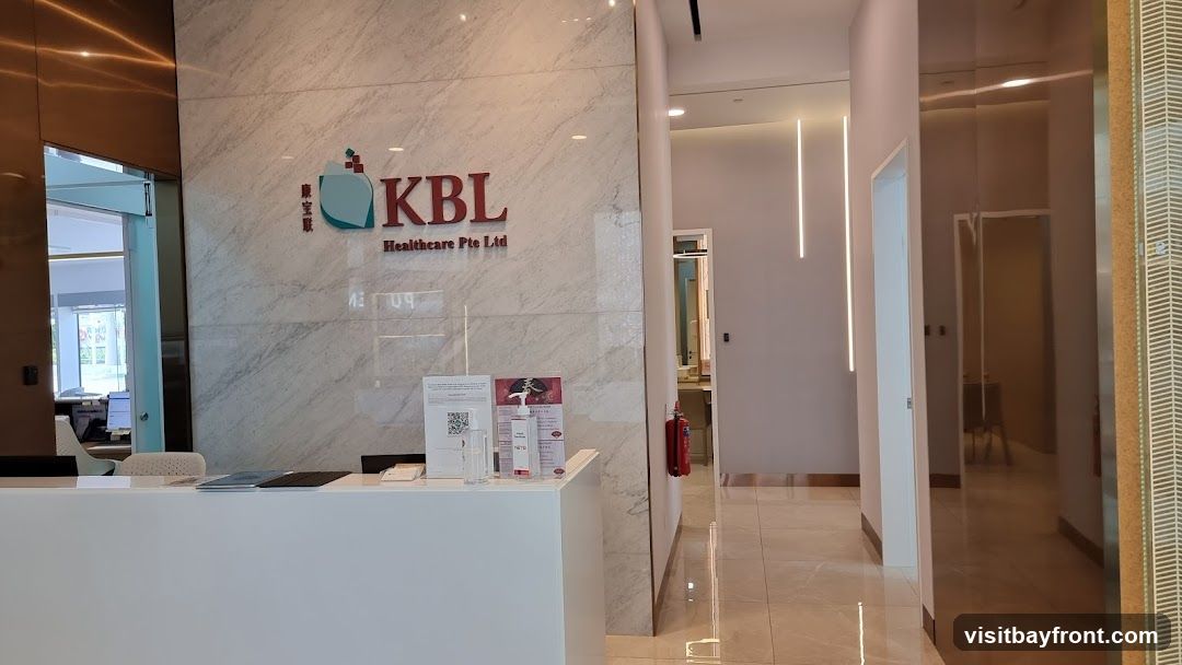 A modern reception area with a white counter and a marble wall featuring the KBL Healthcare Pte Ltd logo. There are informational flyers and hand sanitizer on the counter. A hallway leads to the right, with a fire extinguisher visible.