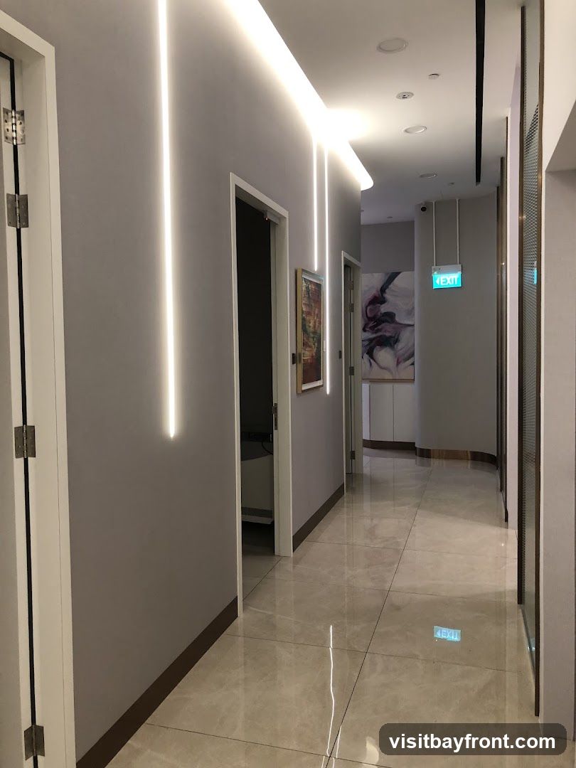 A clean and modern hallway with grey walls and marble-tiled floors. Vertical LED strip lights are embedded in the wall, creating a sleek and contemporary ambiance. Several doors line the hallway, some open to reveal glimpses of rooms. Artwork hangs on the walls, adding a touch of sophistication. An illuminated green 'EXIT' sign is visible in the distance, indicating a safety feature. The overall impression is one of a well-maintained and stylish interior space, possibly a clinic, spa, or office building.