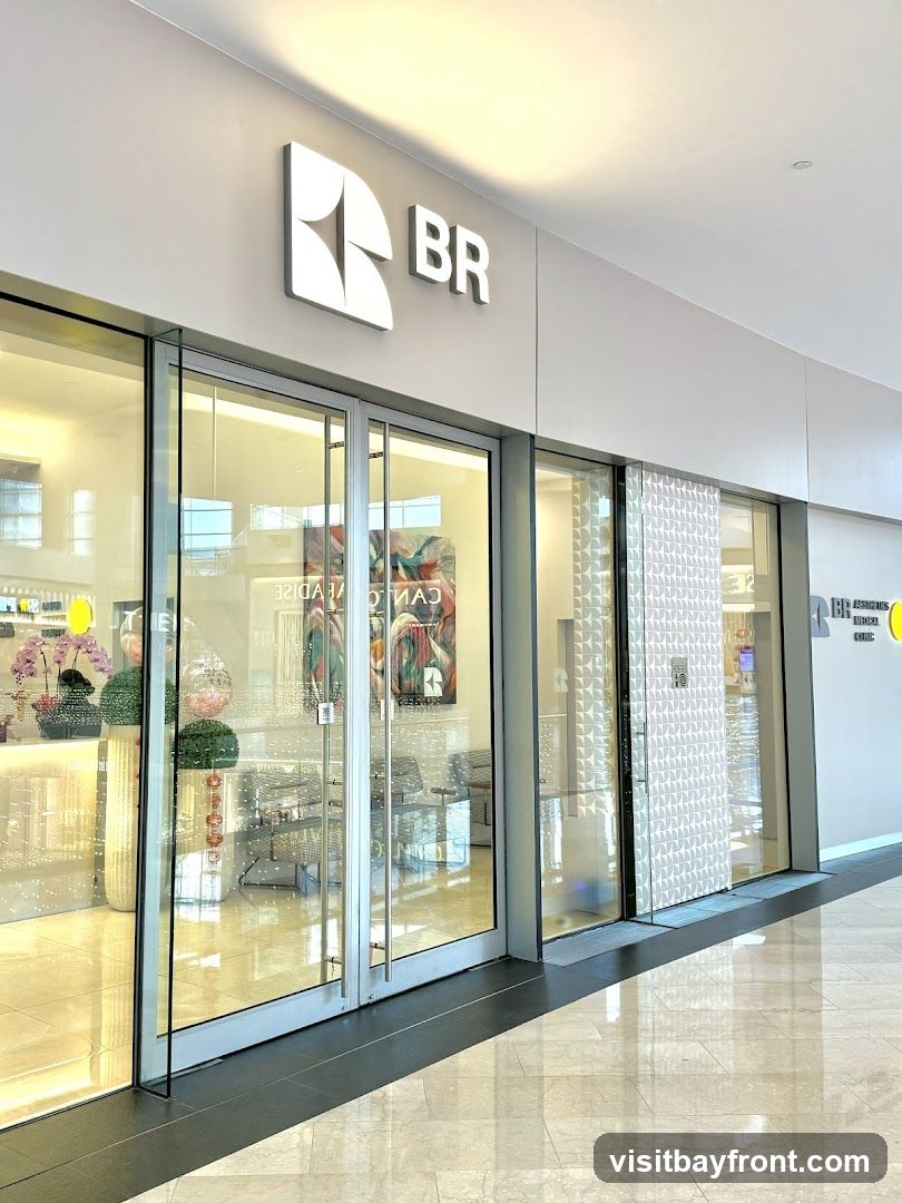 The exterior of a modern medical clinic with large glass windows and doors. A prominent white logo and the letters "BR" are displayed above the entrance. Inside, the clinic appears to be well-lit and features decorative elements like plants and artwork.