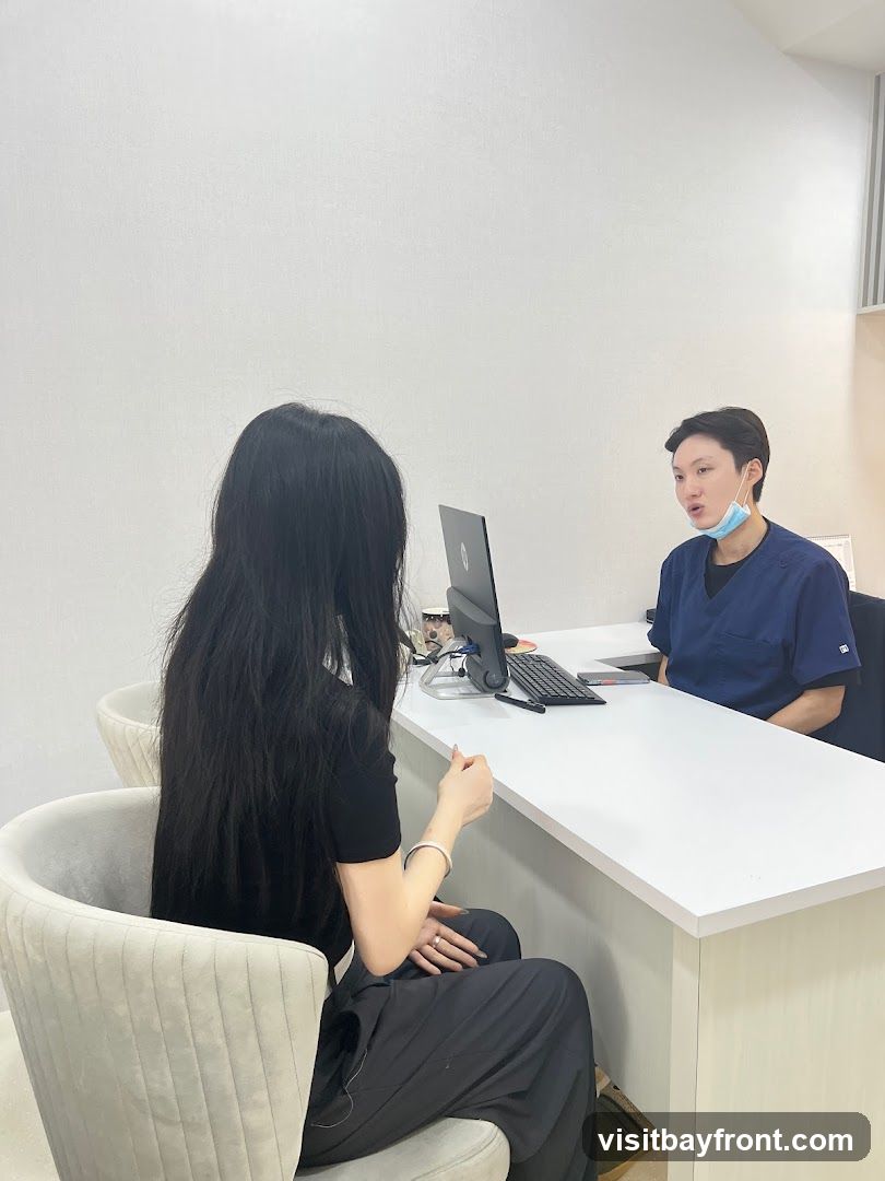 A person with long black hair sits in a chair facing a desk, while another person in blue scrubs, wearing a face mask, sits behind the desk and speaks. A laptop and keyboard are on the desk.
