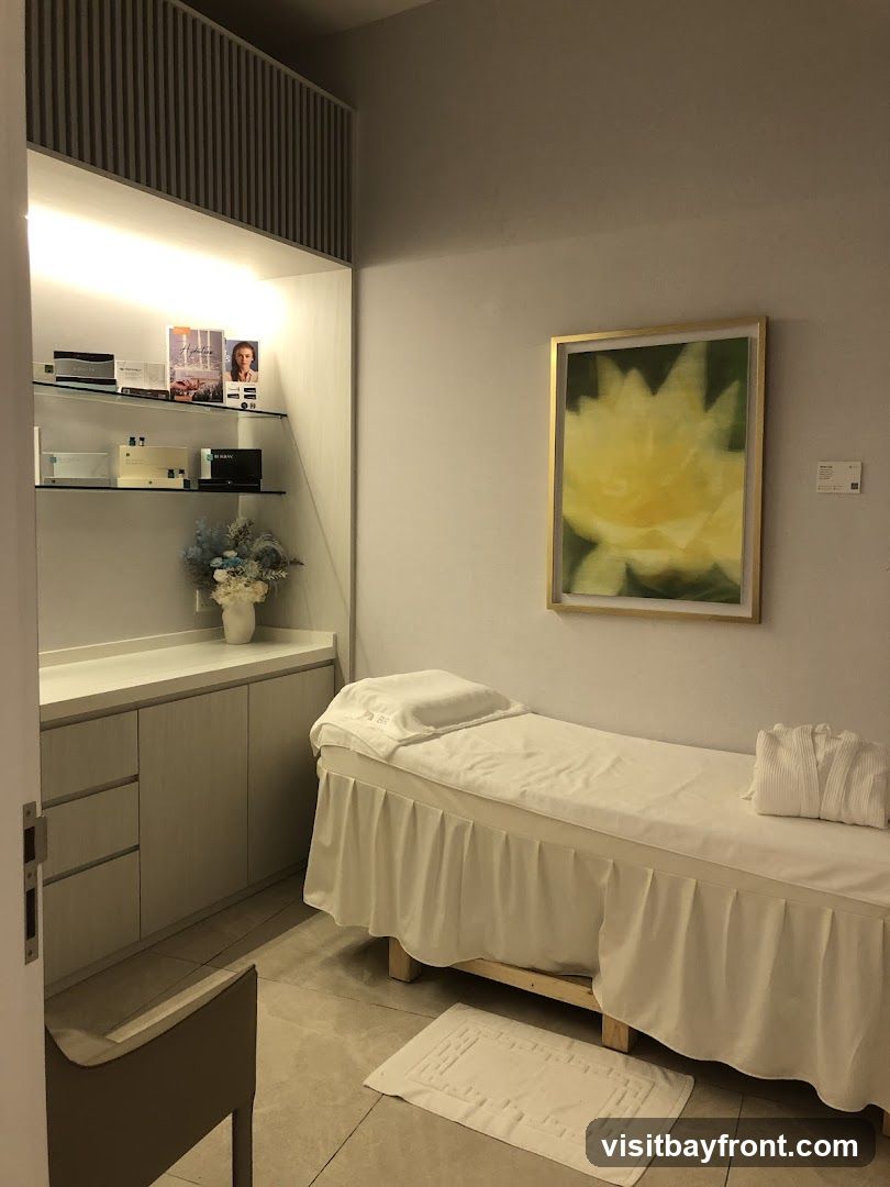 A clean and serene spa treatment room with a massage bed covered in white linens, a folded white robe, and a small rug on the floor. Shelves on the left display various skincare products and brochures. A framed artwork of a yellow flower hangs on the wall.