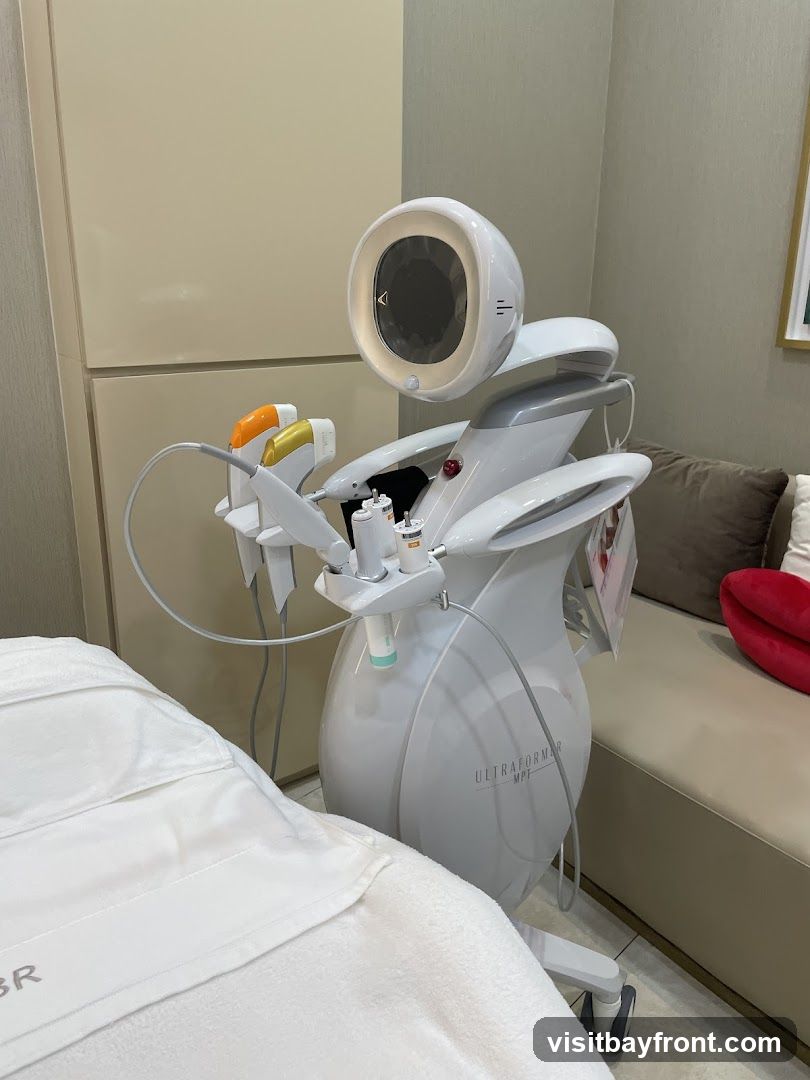 A white medical aesthetic device with a large circular screen and multiple handpieces is positioned next to a treatment bed covered in white linens. The device has the branding "ULTRAFORMER MPT" visible on its side. The room appears to be a clean, modern clinic or spa setting with beige walls and cabinetry, and a cushioned bench in the background.