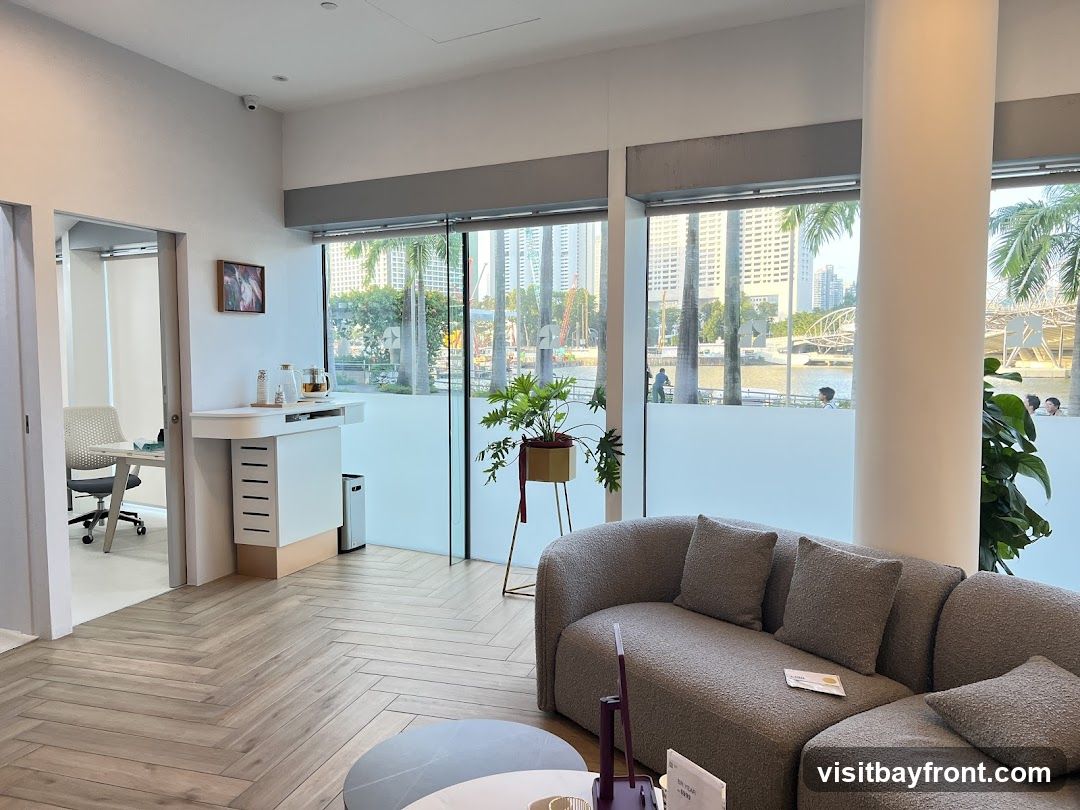 A comfortable and modern waiting area with a plush grey sectional sofa, a round coffee table, and a plant in a gold pot. Large windows offer a view of a city skyline, a river, and palm trees. To the left, a doorway leads to an office space with a desk and chair. A white counter with a kettle and bottles is also visible.