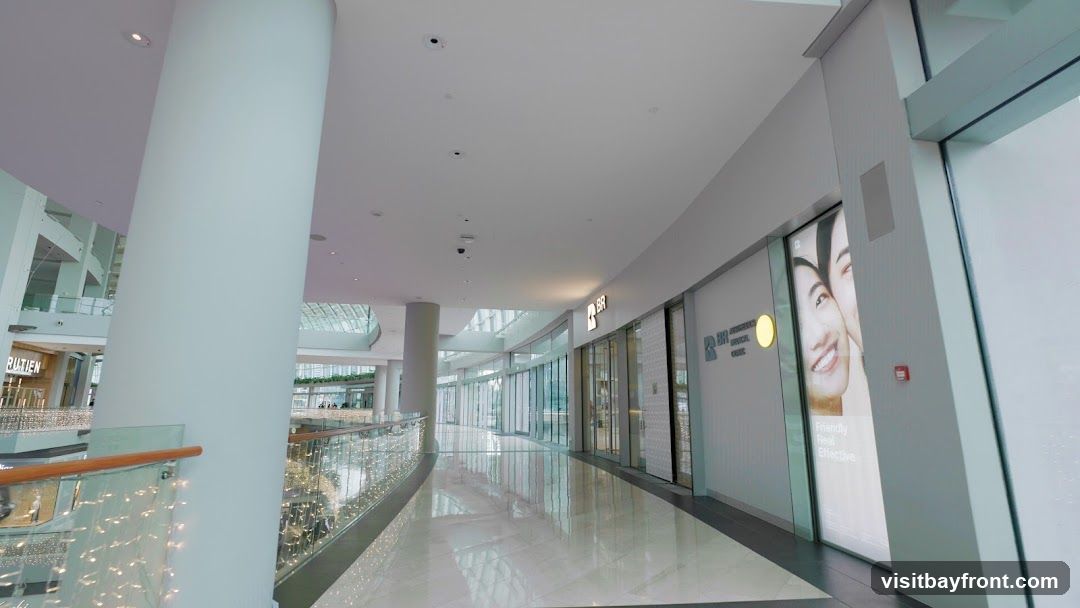 A wide shot of a modern, well-lit shopping mall interior. A large white pillar is on the left, with a railing decorated with fairy lights. The walkway is polished and reflects the ceiling lights. On the right, several storefronts are visible. One storefront has a large advertisement featuring two smiling faces and text that reads "Friendly Real Effective." Another storefront displays the logo "BR" and appears to be a clinic. In the distance, more levels of the mall are visible, with shops and escalators.
