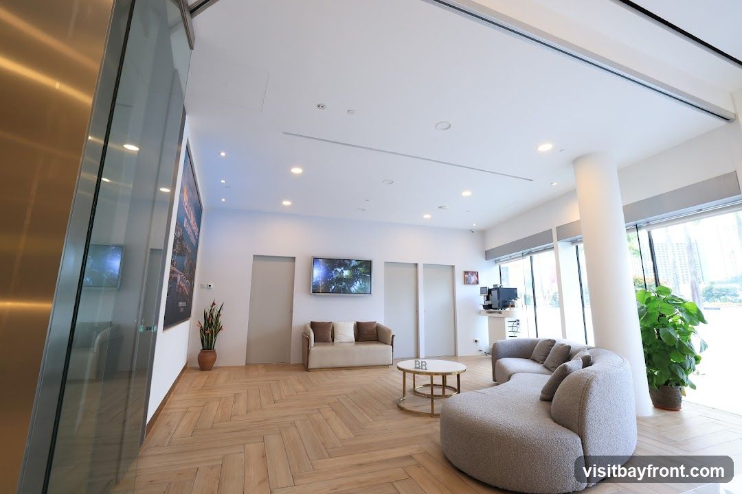 A contemporary waiting area features a large, curved sofa and a smaller sofa, both with plush cushions. A round coffee table with a metallic base sits between them. On the wall, a large framed artwork is displayed, and two flat-screen televisions show nature scenes. The floor is laid with light-colored wooden planks in a herringbone pattern. A coffee machine is visible on a counter near a large window overlooking a cityscape. A tall potted plant adds a touch of greenery to the space.