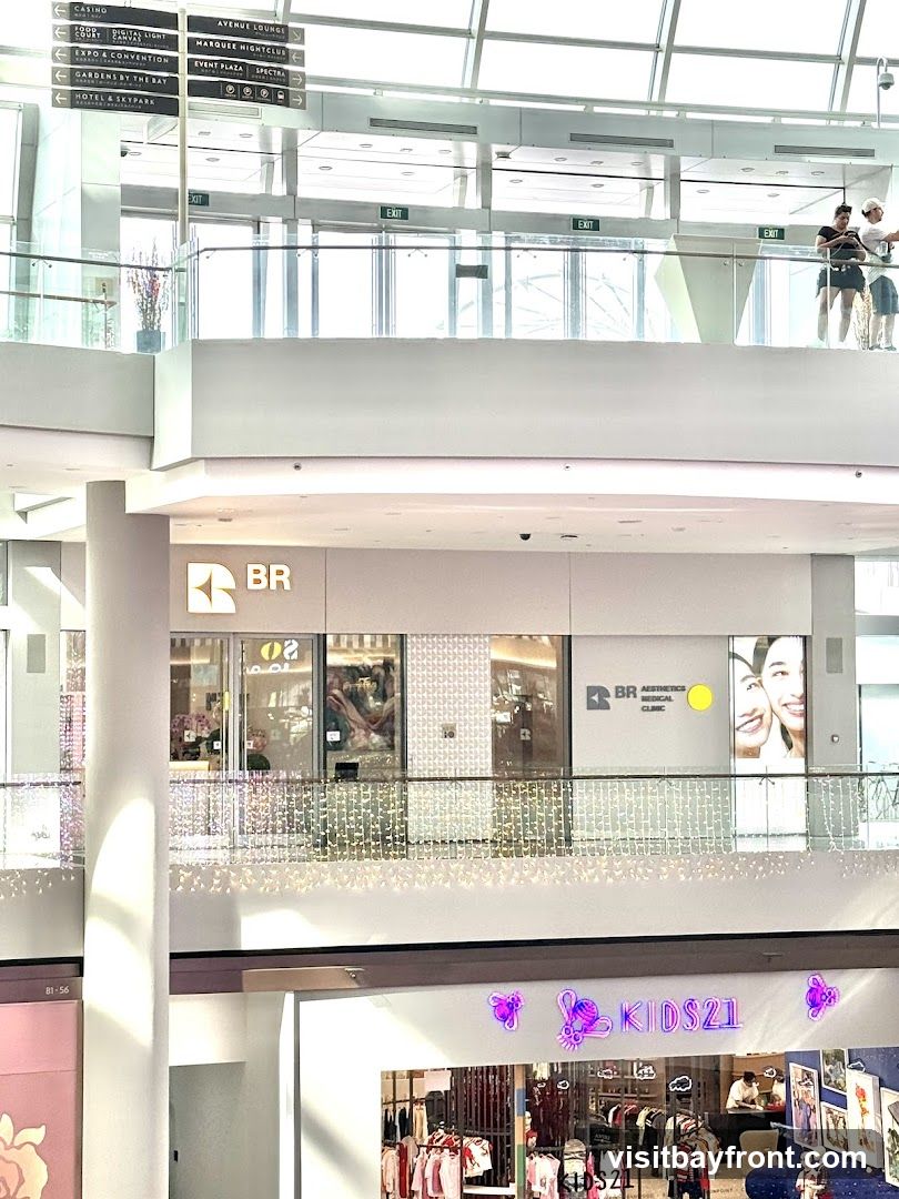 An interior view of a multi-level shopping mall. On the upper level, signage indicates various locations like