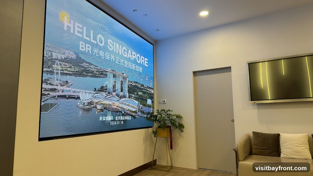 A large digital screen displays a promotional image of Singapore landmarks, including the Marina Bay Sands, with the text "HELLO SINGAPORE" and "BR 光电保养正式登陆新加坡" (BR Optoelectronics officially lands in Singapore). Below, it reads "新加坡BR·金沙酒店体验店" (Singapore BR·Marina Bay Sands Hotel Experience Store) and the date "2024.01.18". The screen is mounted on a wall in a room with a closed door, a sofa with cushions, and a potted plant. A smaller screen with yellow vertical lines is mounted on the opposite wall.