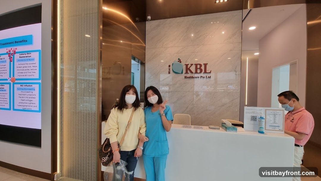 Two women and a man are at the reception desk of KBL Healthcare Pte Ltd. One woman is wearing a yellow sweater and the other is wearing a blue scrub suit. The man is wearing a pink polo shirt. There is a digital display on the left showing treatment benefits. The reception desk has hand sanitizer and some papers. The KBL Healthcare logo is visible on the wall behind the reception desk.