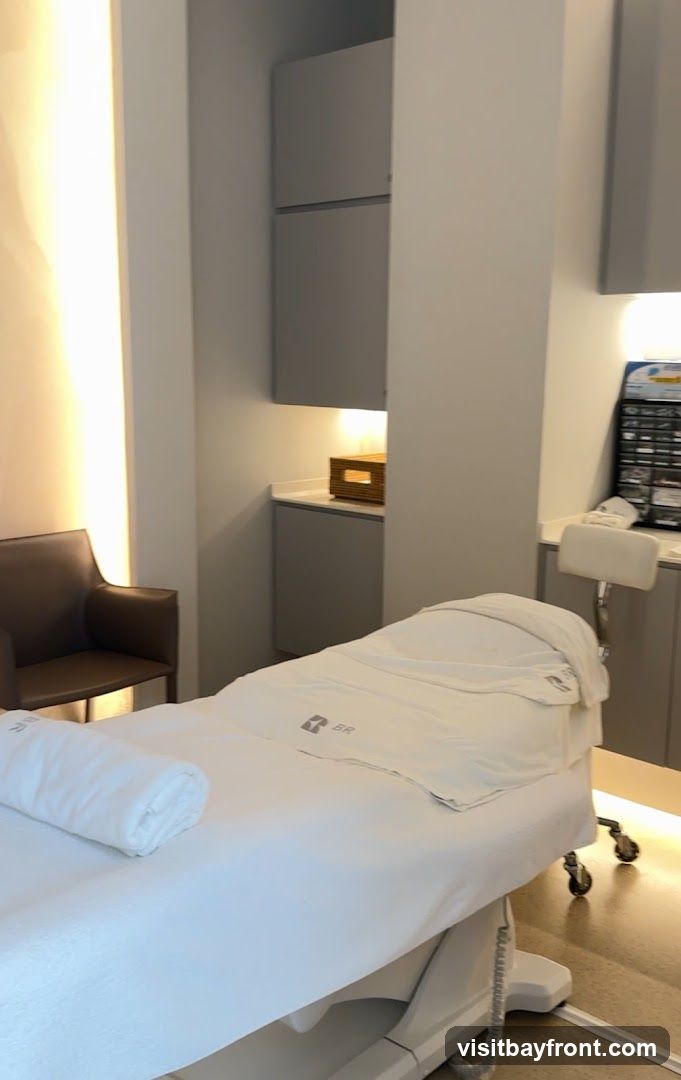 A clean and modern spa treatment room with a massage bed covered in white linens, a rolled towel, and a logo. There is a brown leather chair to the left and grey cabinets and shelving in the background. Soft lighting illuminates the space.