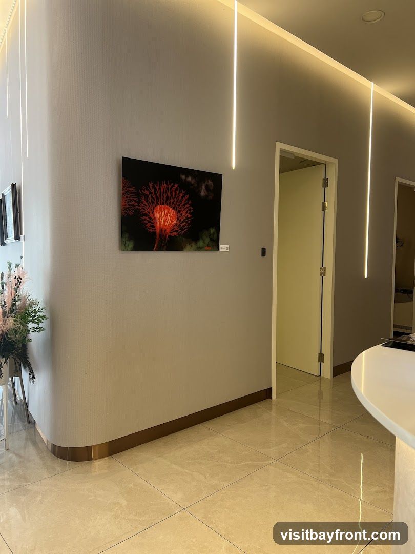 A contemporary interior space featuring a large abstract artwork of a red, spiky flower on a grey textured wall. Vertical LED strip lights illuminate the ceiling and walls. A doorway with a glass door is visible on the right, leading to another room. A curved reception desk or counter is partially visible in the foreground. The floor is tiled with a light-colored marble pattern. A floral arrangement is placed in the corner to the left.