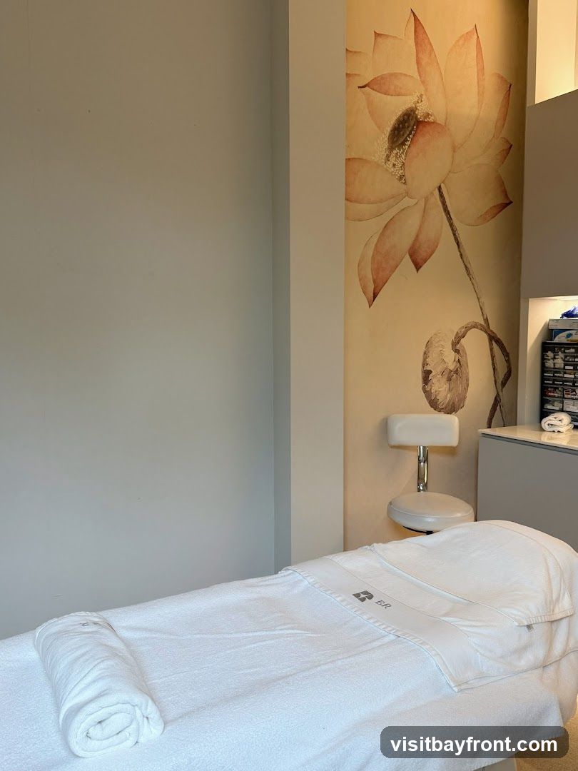 A spa room with a massage table covered in white linens and a rolled towel. A floral mural adorns the wall behind a white stool and a cabinet with supplies.