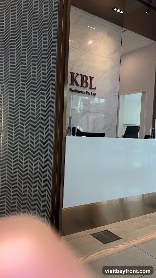 A view of the entrance to KBL Healthcare Pte Ltd, featuring a white reception desk and marble-patterned walls. The company logo, 'KBL Healthcare Pte Ltd', is prominently displayed on the wall behind the desk. The entrance has a modern and clean aesthetic.