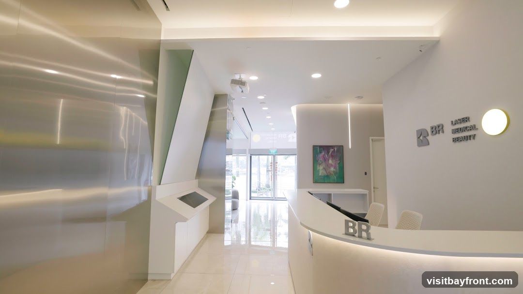 A sleek and modern reception area with a white curved counter. The wall behind the counter features a logo that reads "BR LASER MEDICAL BEAUTY" and a circular light fixture. A contemporary painting hangs on the wall. To the left, a wall with a brushed metal finish leads to a hallway with an exit sign visible through glass doors. There are interactive touch screens on a white console.