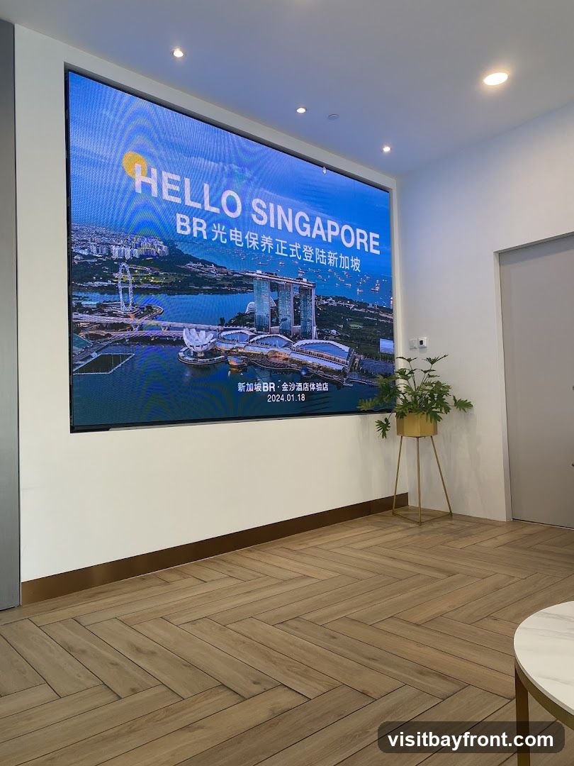 A large digital screen displays "HELLO SINGAPORE" with an aerial view of the Singapore skyline, featuring the Marina Bay Sands hotel and the Singapore Flyer. Below the main text, it reads "BR光电保养正式登陆新加坡" (BR Optoelectronics officially lands in Singapore) and "新加坡BR·金沙酒店体验店" (Singapore BR · Marina Bay Sands Hotel Experience Store), with the date "2024.01.18". The screen is mounted on a white wall above a herringbone-patterned wooden floor. A potted plant in a gold stand is visible to the right of the screen.